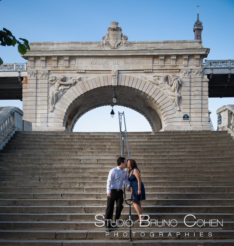 portrait kissing couple in love from Bir Hakeim bridge stand front arche at sunrise proposal in paris blue sky