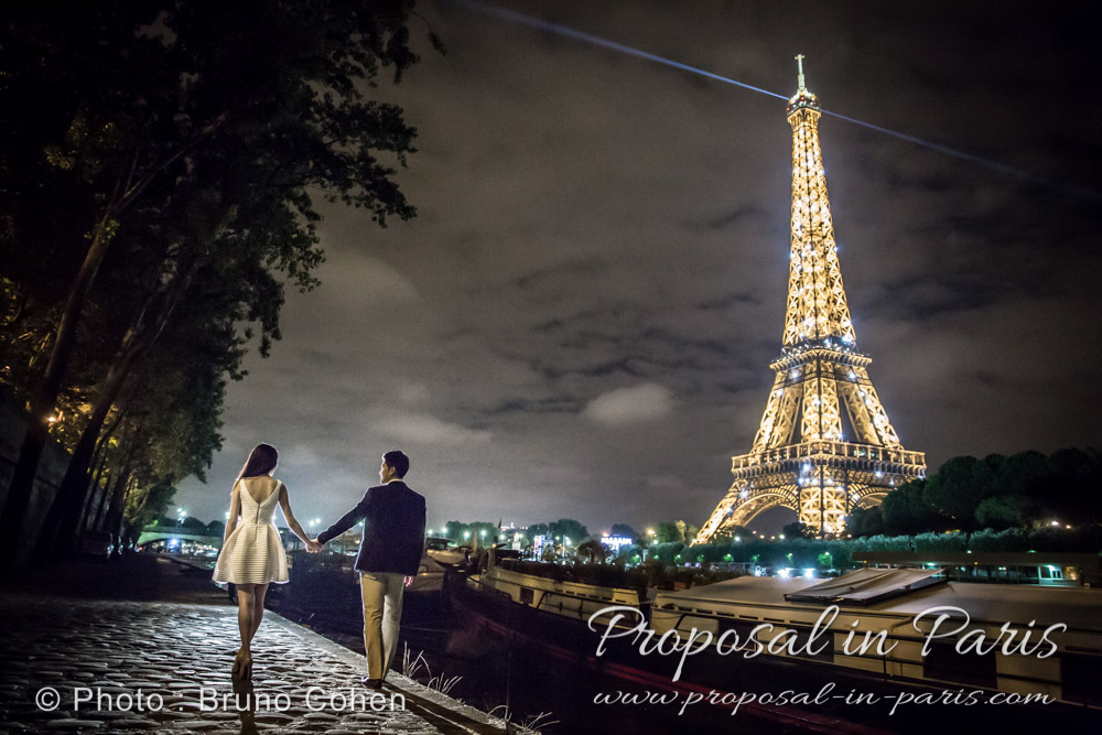 a couple is walking along the Seine river by night near the Eiffel Tower